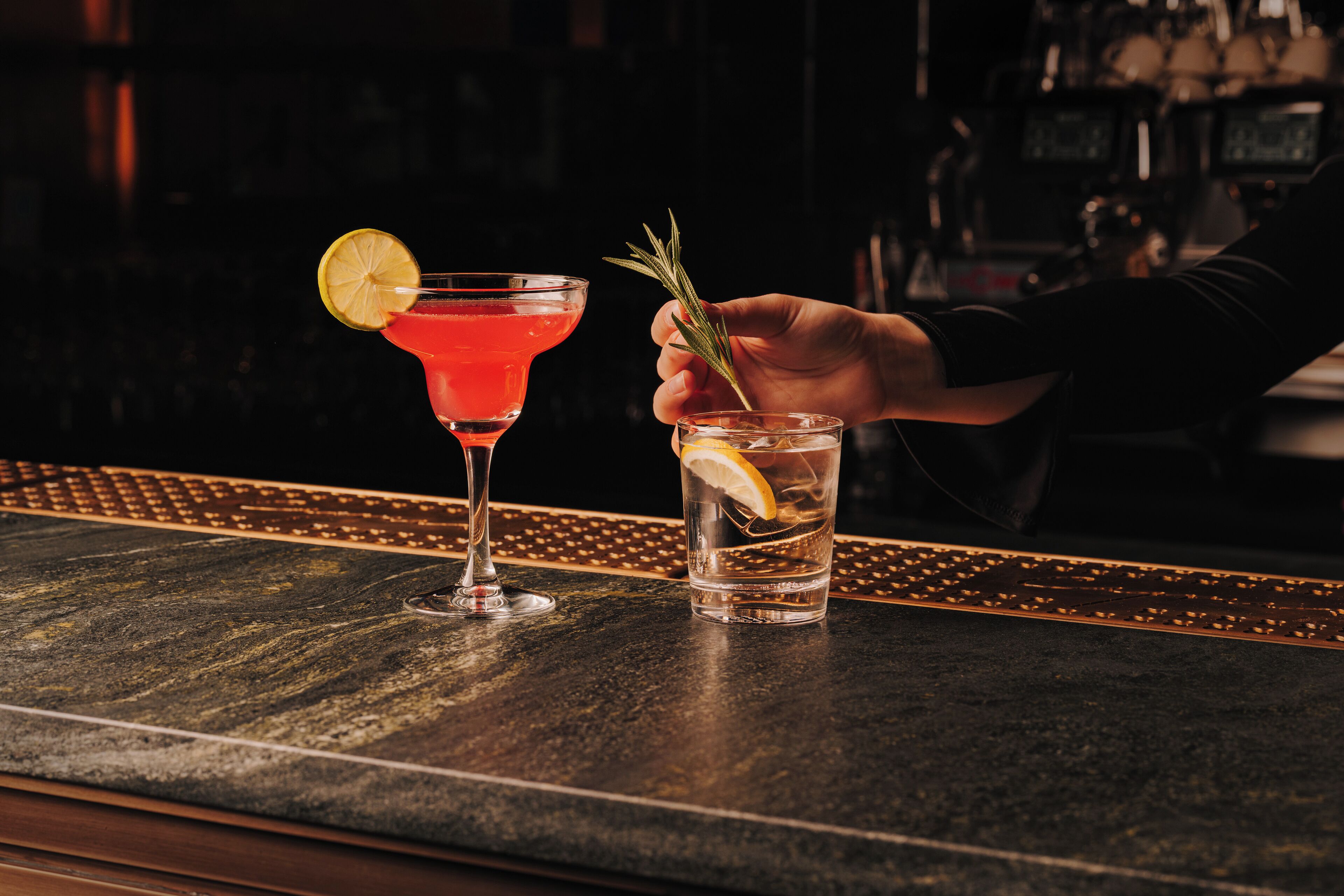 A bar counter with a martini glass and a clear glass with a lemon slice and a sprig of rosemary.