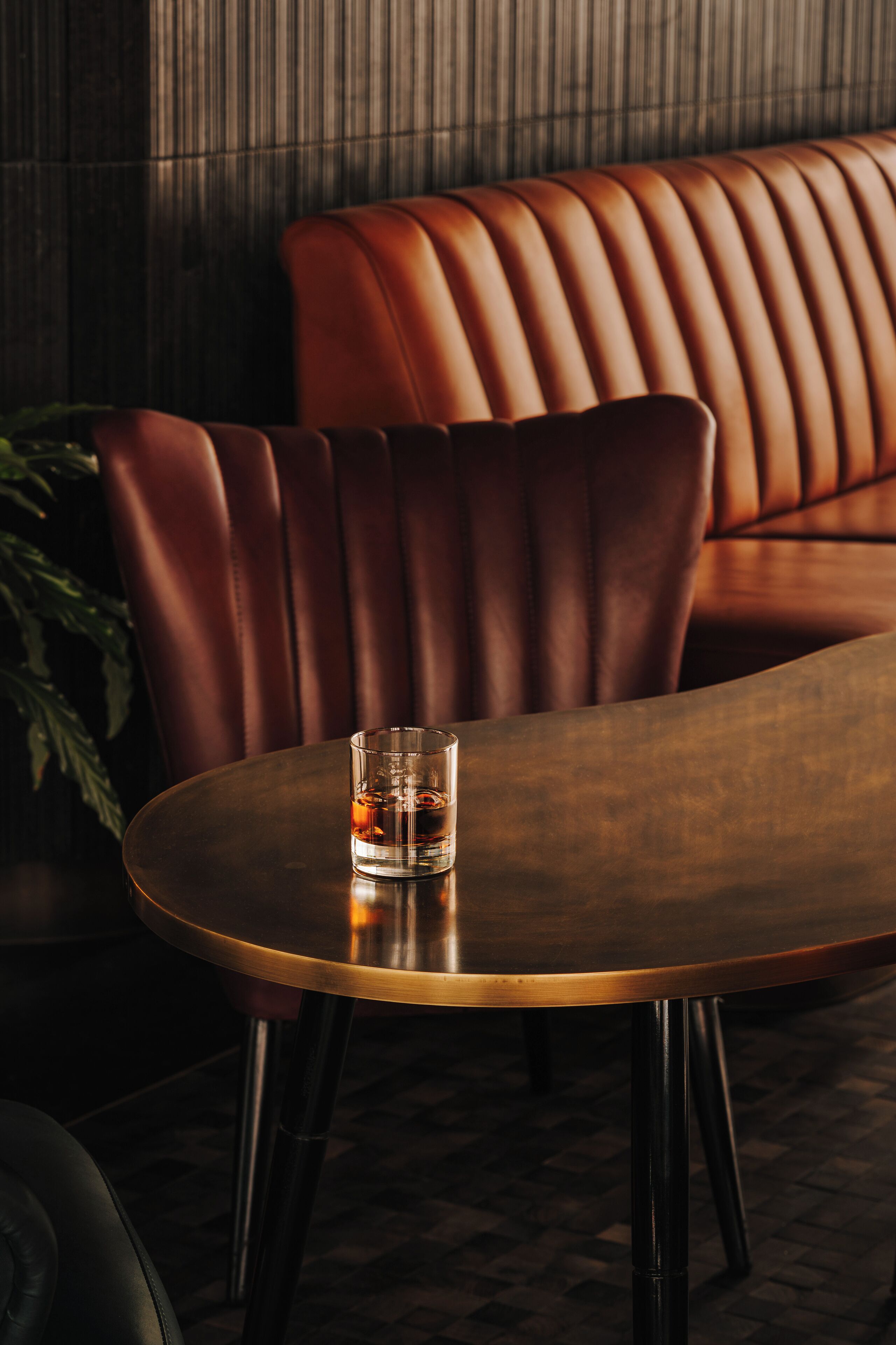 A round table with a glass of whiskey, a brown leather couch, and a plant in a dimly lit room.