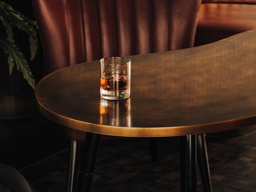 A round table with a glass of whiskey, a brown leather couch, and a plant in a dimly lit room.