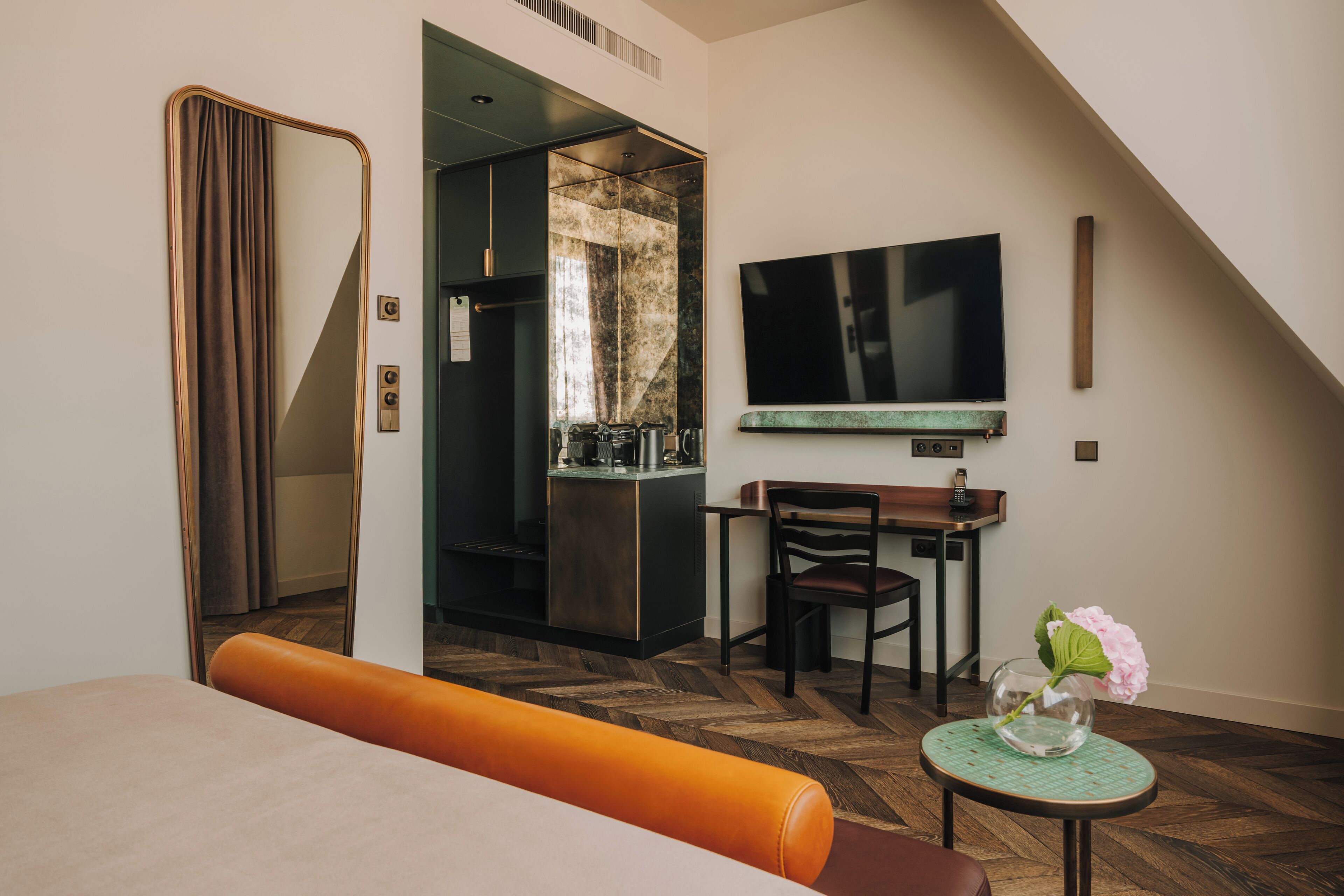 Une chambre d’hôtel moderne dotée d’un grand miroir, d’un téléviseur à écran plat, d’un coin repas et d’une petite table sur laquelle est posé un vase de fleurs.