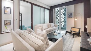 Una sala de estar moderna con un sofá, una mesa de centro y un escritorio con una lámpara. Hay un ventanal corredero que da a un balcón con cortinas estampadas.