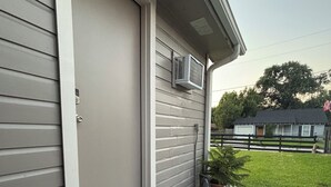 A house exterior with a grey siding, a closed door, a small air conditioning unit mounted on the wall, a wooden bench outside, and a paved area with stepping stones.
