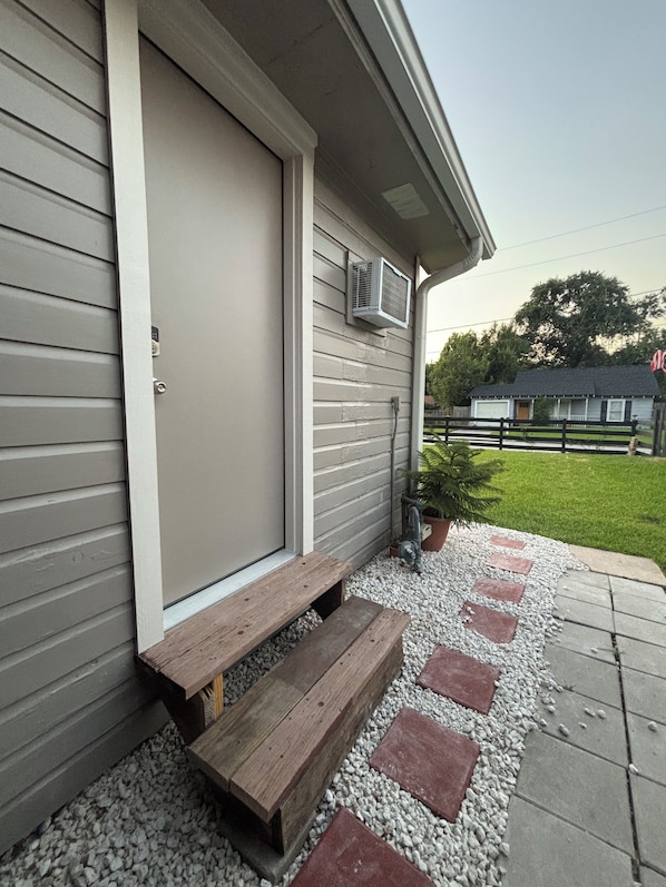 A house exterior with a grey siding, a closed door, a small air conditioning unit mounted on the wall, a wooden bench outside, and a paved area with stepping stones.
