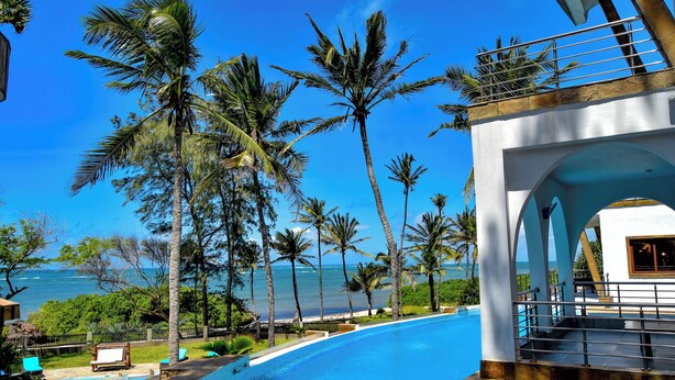 Zona de piscina con vista al océano, rodeada de palmeras y un edificio blanco con balcón.