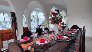 Comedor con una larga mesa dispuesta para una comida, con vista al océano a través de ventanas arqueadas.