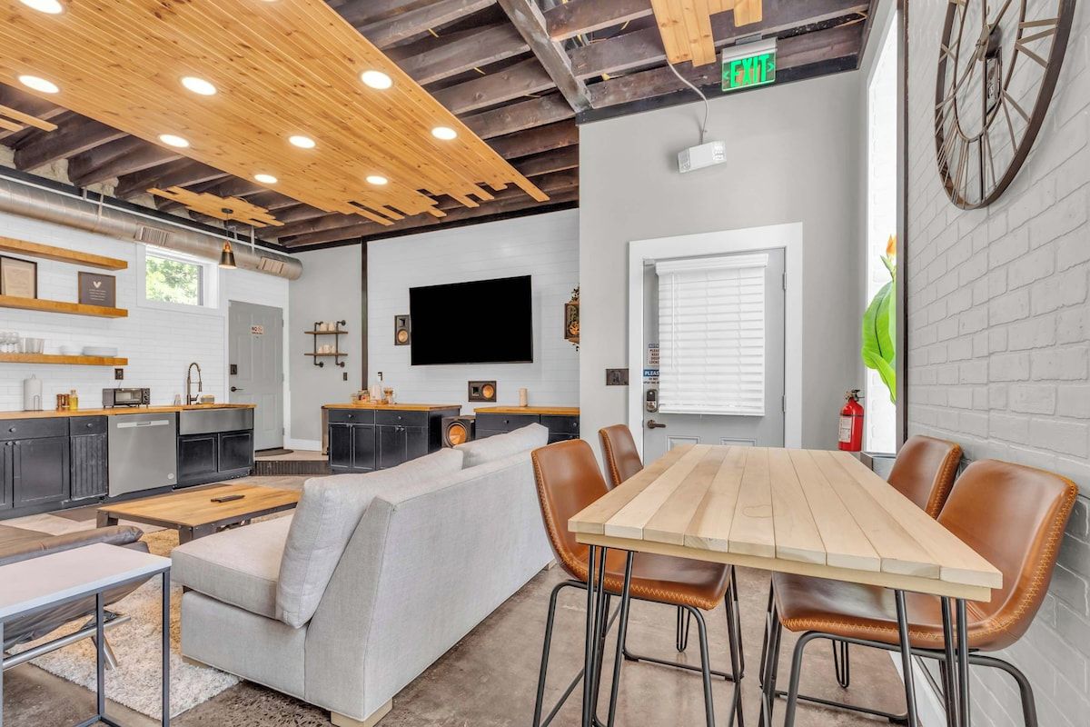 Une cuisine moderne dotée d’un four encastré, un coin repas avec une table en bois et des chaises en cuir, et une grande horloge au mur.