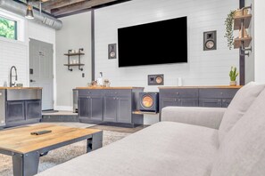 A modern living room with a large flat-screen TV mounted on the wall, a sofa, a coffee table, and a kitchen area with grey cabinets and a wooden countertop.