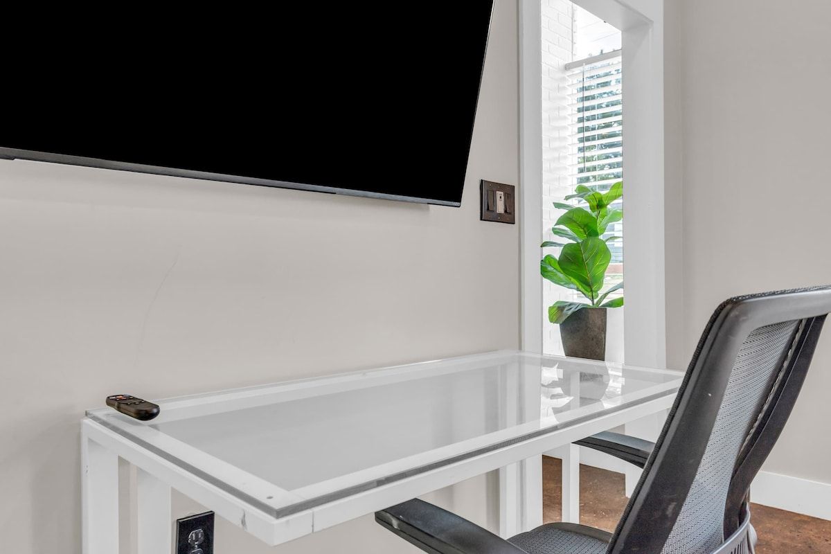 A room with a white desk, a black chair, a mounted television, and a window with blinds.