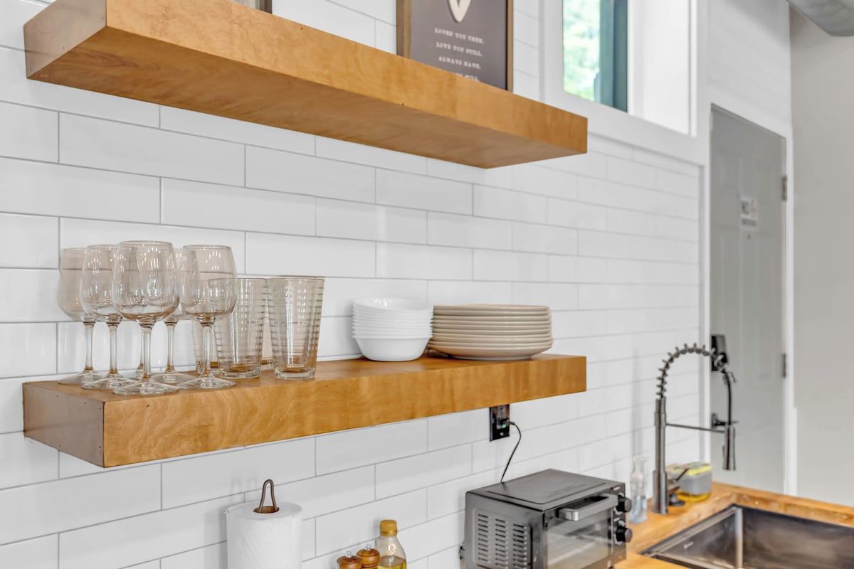A kitchen with white tiled walls, a wooden shelf holding glasses and plates, a roll of paper towels, and a microwave.