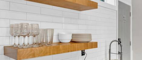 A kitchen with white tiled walls, a wooden shelf holding glasses and plates, a roll of paper towels, and a microwave.