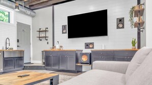 A modern living room with a large flat-screen TV mounted on the wall, a grey sofa, a wooden coffee table, and a kitchen area with grey cabinets and a built-in washer. - Walk to Downtown, King Bed, great for long stays (Spartanburg)