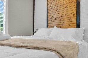 A bedroom with a bed, white bedding, a wooden headboard, and a window with blinds. - Industrial Loft - Walk to Downtown, Patio w/ BBQ (Spartanburg)