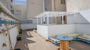 A small outdoor patio area with a wooden table, a white bench, and a small covered seating area.