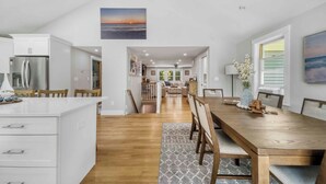 A modern kitchen with white cabinets and a large island, a wooden dining table with chairs, and a view of the living area through an open archway. - Recently renovated 3 bedroom (Falmouth)