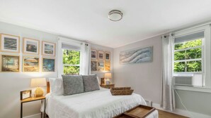 A bedroom with a bed, bedside tables, a nightstand, a lamp, a picture frame, a wall-mounted air conditioner, and a window with curtains. - Recently renovated 3 bedroom (Falmouth)