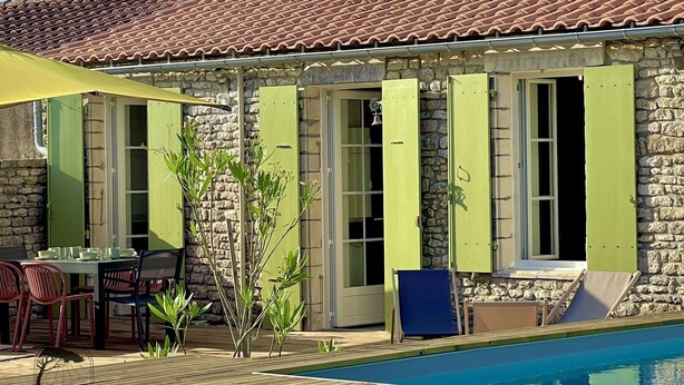 Un espace aménagé au bord de la piscine, avec un bâtiment en pierre, des volets verts et du mobilier d’extérieur.