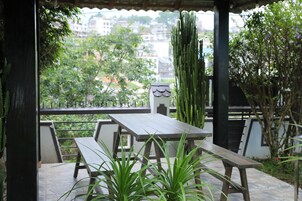 Alpendre coberto com mesa e bancos de madeira, rodeado por plantas em vasos e vista para edifícios e árvores.
