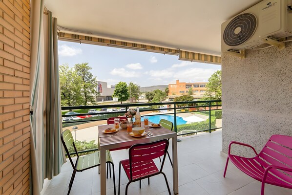 A balcony with a table set for two, a chair, a fan, and a view of a swimming pool and buildings.
