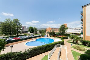 A swimming pool surrounded by greenery, a parking lot, and buildings in the background.