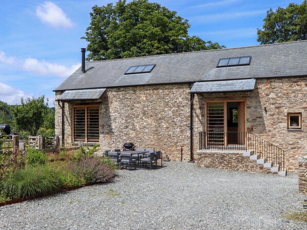 Un bâtiment en pierre avec une allée de gravier, un espace barbecue et un escalier menant à une porte en bois.