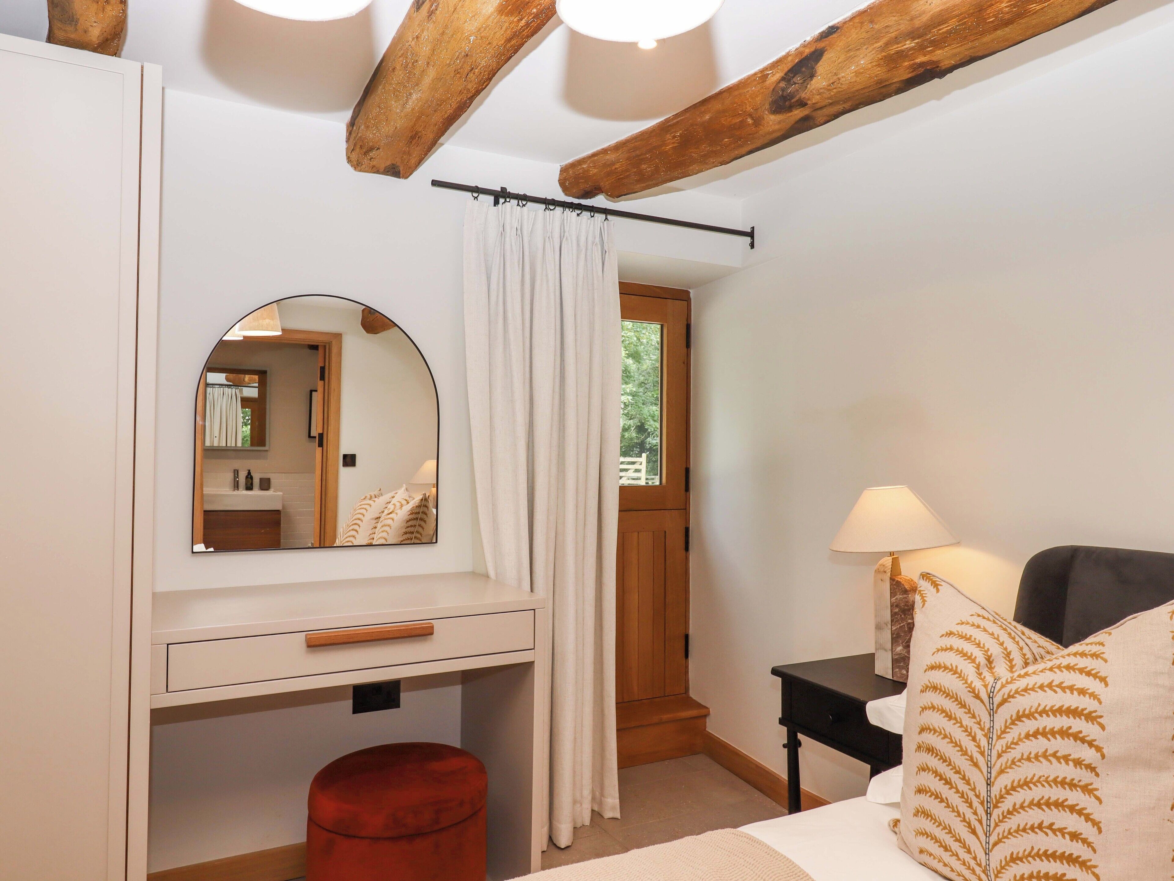 Un dormitorio con techo de madera, un espejo, una mesita de noche con lámpara, un otoman rojo y una puerta que da a otra habitación.