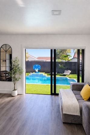 A living room with a grey sofa, a potted plant, and a view of a backyard with a pool and a basketball hoop.