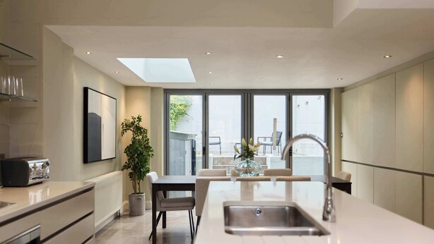 A modern kitchen with a central island, a dining area with chairs, a potted plant, and a view through large windows. - Redbrick on Langton (London)