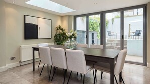 A dining area with a table, chairs, a vase with flowers, a painting on the wall, and a view through large windows. - Redbrick on Langton (London)