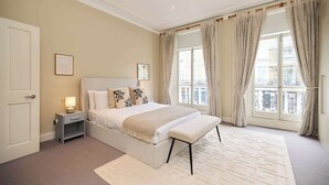 A bedroom with a bed, bedside table, bench, and large window with curtains. - Redbrick on Langton (London)