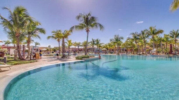 Une grande piscine à eau claire, entourée de palmiers et de chaises longues, sous un ciel dégagé.