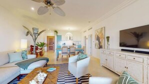 Un salon moderne avec un canapé, des fauteuils, une table basse et un téléviseur. On aperçoit à l’arrière-plan un coin repas avec une table et des chaises.