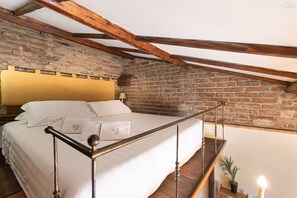 Una camera da letto con parete in mattoni, trave in legno al soffitto, un letto con lenzuola bianche e un comodino con lampada.