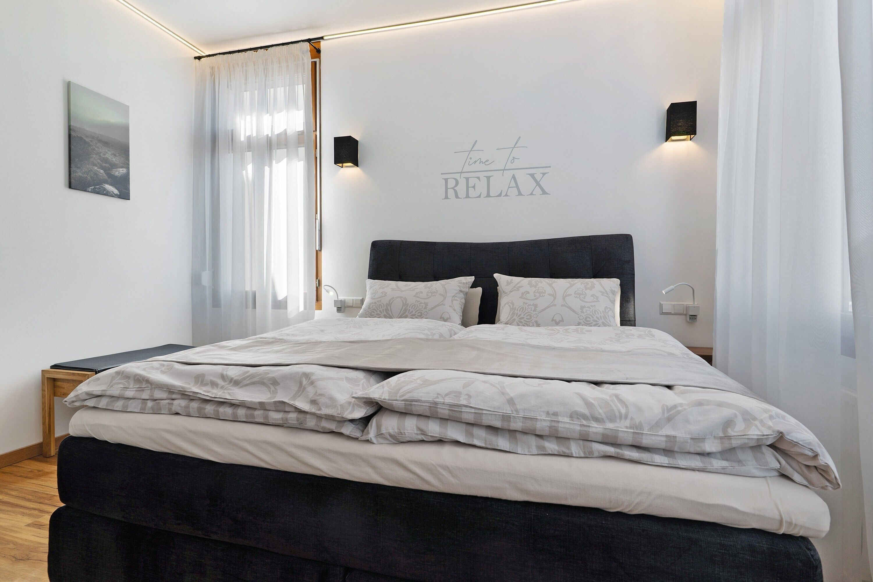 En pænt redt seng med gavl, et natbord, en vægmonteret lampe og et skilt med teksten 'time to RELAX'.