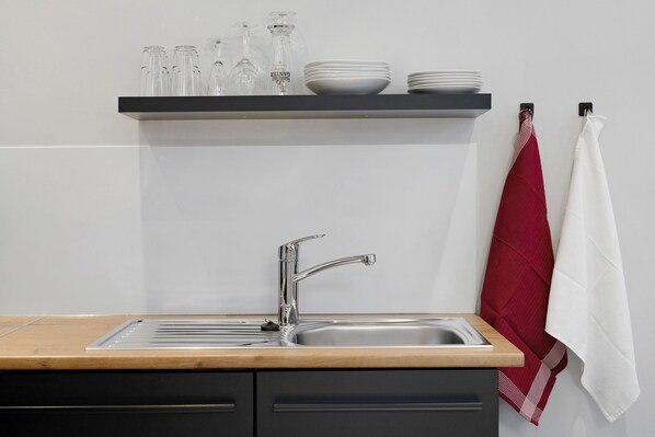 A kitchen with a stainless steel sink, wooden countertop, and a shelf above holding glassware and plates.
