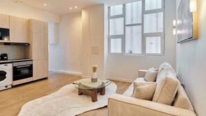 A modern living room with a glass coffee table, beige sofa, and a large window with blinds. - Elegant Apartment With Buttes-chaumont Park (Paris)
