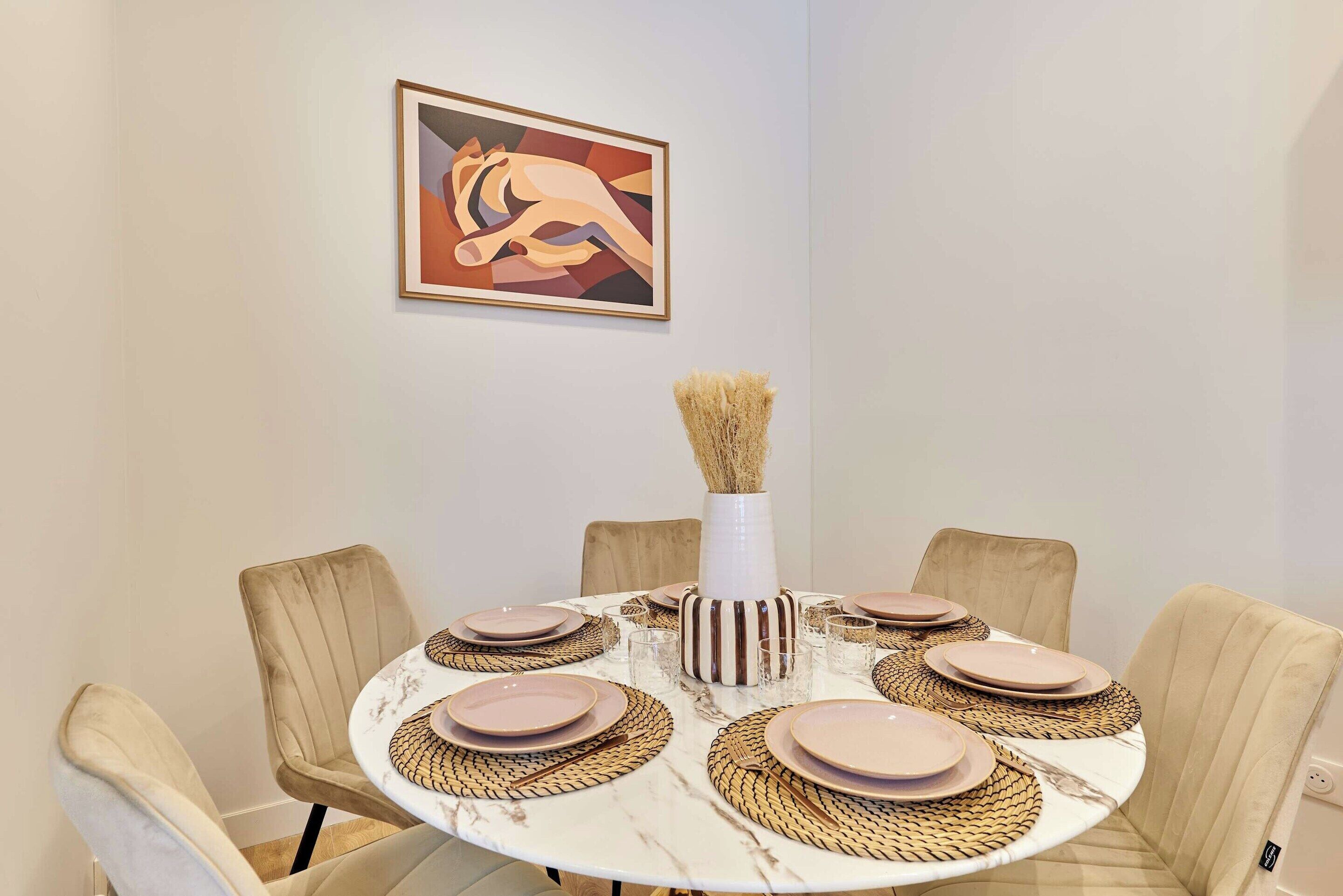 Une table de salle à manger ronde, avec des chaises recouvertes d’un tissu beige, un vase blanc contenant des plantes séchées et, au mur, un tableau abstrait encadré.