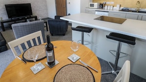 A modern kitchen with a central island, bar stools, a dining table with a bottle of wine, and a TV in the background. - Quayside Apartments (Whitehaven)