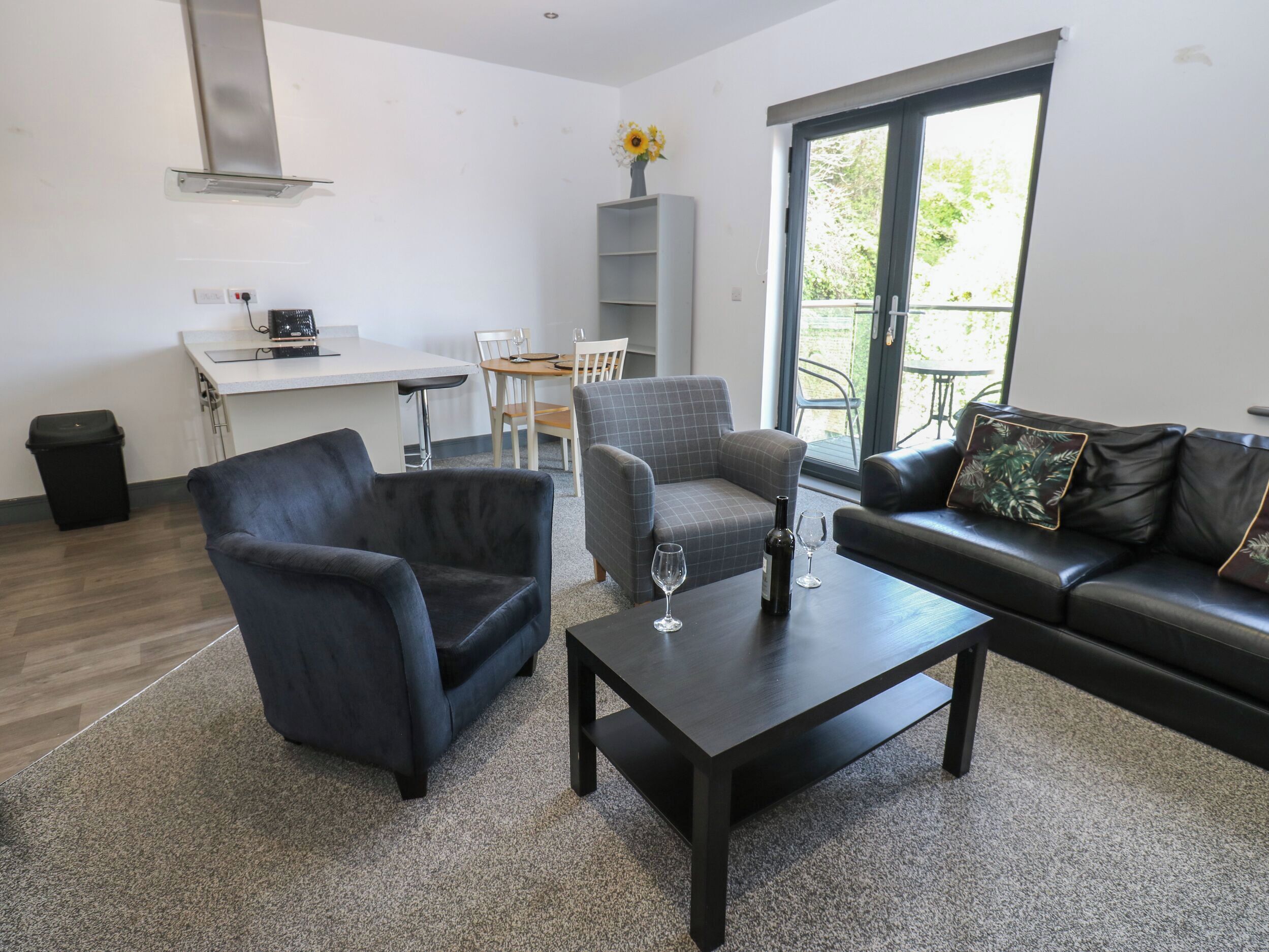 A modern living room with a dark leather sofa, a grey armchair, and a black coffee table. There is a dining area with a white table and chairs, and a kitchen area with a white countertop and a built-in stove.