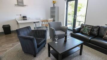 A modern living room with a dark leather sofa, a grey armchair, and a black coffee table. There is a dining area with a white table and chairs, and a kitchen area with a white countertop and a built-in stove.