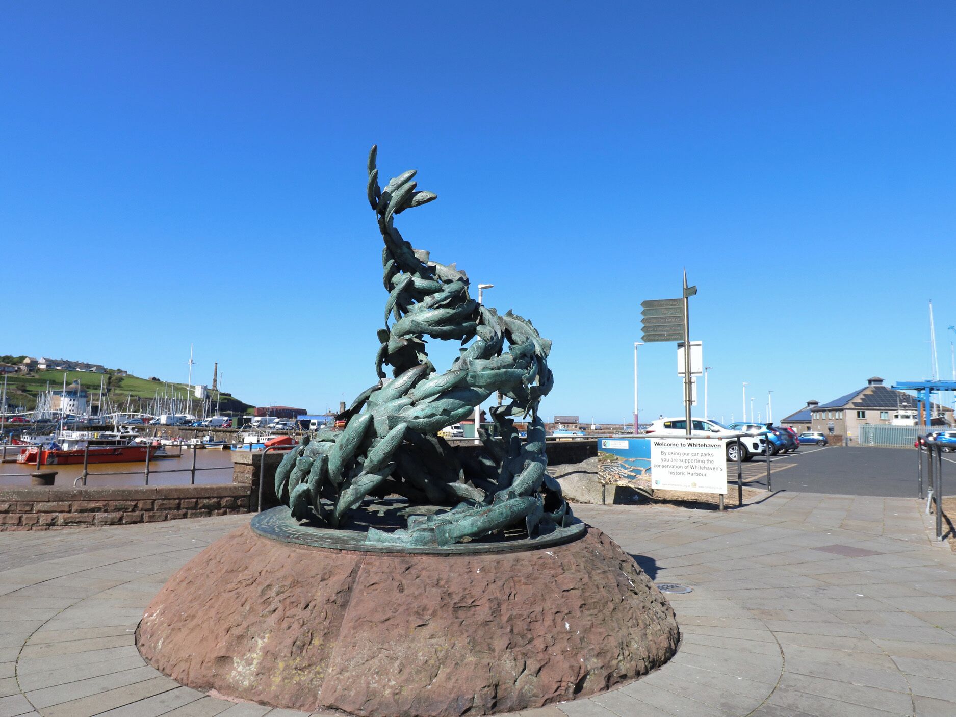 Una escultura en una plaza pública, cielo despejado y barcos al fondo.