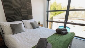 A neatly made bed with a green towel and two rolled towels, a window with a view of a building, and a wall with a textured panel design. - Quayside Apartments (Whitehaven)