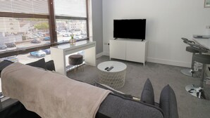 A modern living room with a sofa, a TV on a white cabinet, a round coffee table, and a view of the street through large windows. - Harbourside Apartments (Whitehaven)