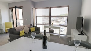 A modern living room with a grey sofa, a dining table with two wine glasses and a bottle, a TV on a stand, and large windows with blinds. - Harbourside Apartments (Whitehaven)