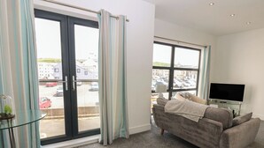 Un salon avec un canapé gris, un téléviseur à écran plat posé sur un meuble en verre, et une vue sur un parking à travers de grandes fenêtres.