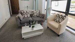 Un salon avec un canapé gris, une table basse blanche et un fauteuil beige. On trouve deux verres à vin et une bouteille sur la table basse. La pièce est dotée d’une grande fenêtre avec des rideaux, d’une porte et d’une petite table avec une lampe.