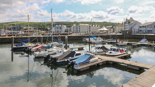 Un port de plaisance où sont amarrés divers bateaux, le tout bordé de quais en bois, et entouré de bâtiments, avec un paysage vallonné en arrière-plan.