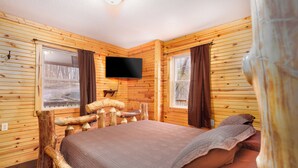 A wooden cabin bedroom with a bed, a mounted TV, and a window with curtains.