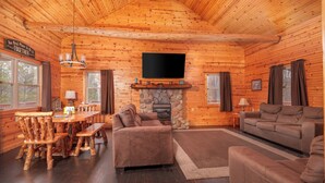 A log cabin interior with a stone fireplace, wooden furniture, and a flat-screen TV mounted on the wall.