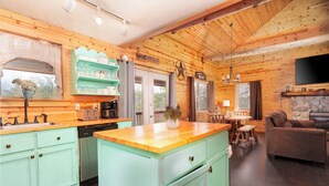 A rustic cabin interior with a kitchen featuring light green cabinetry, a wooden island, and a stone fireplace.
