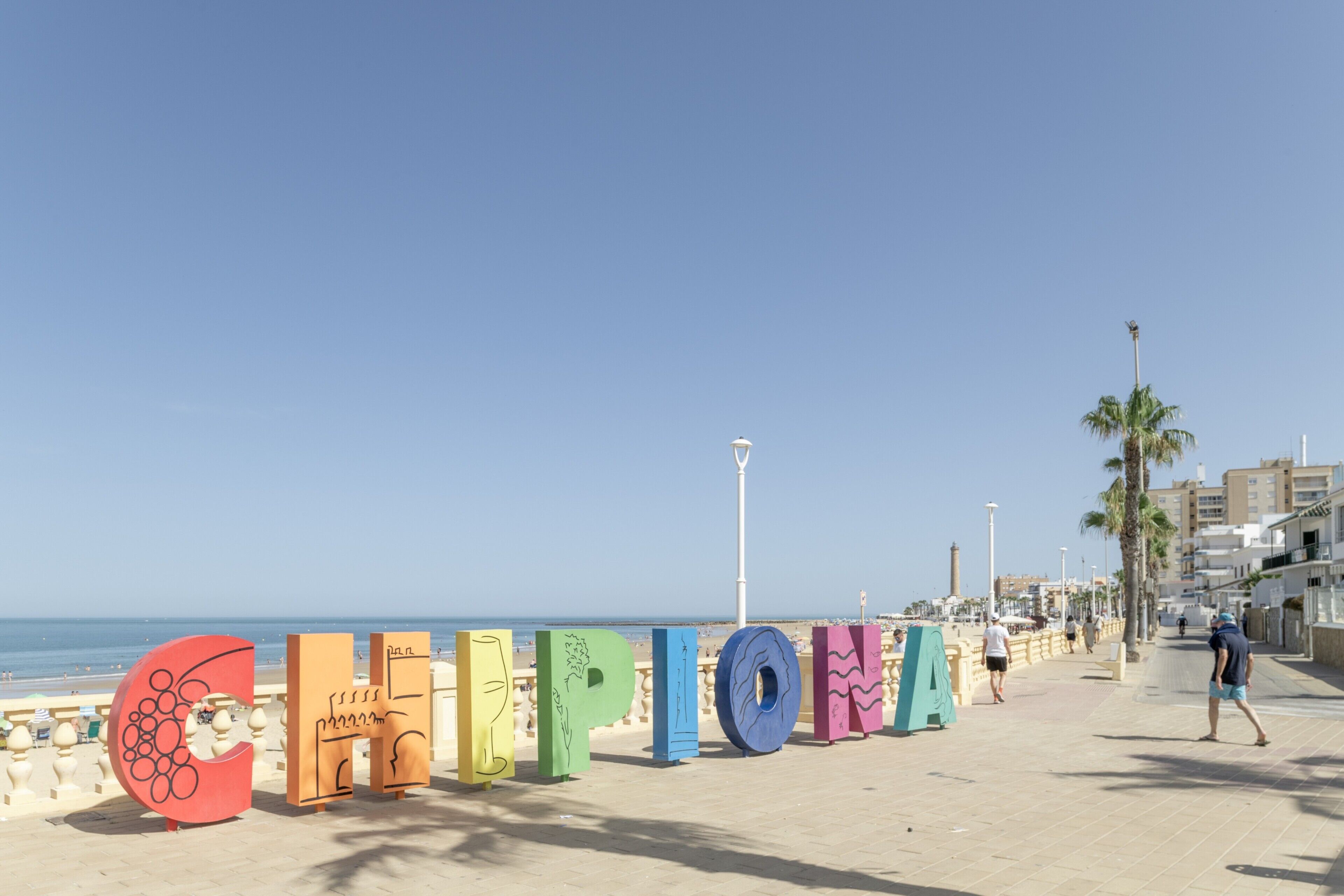 A beachside promenade with large colorful letters spelling 'CHIPIONA' and a clear blue sky.
